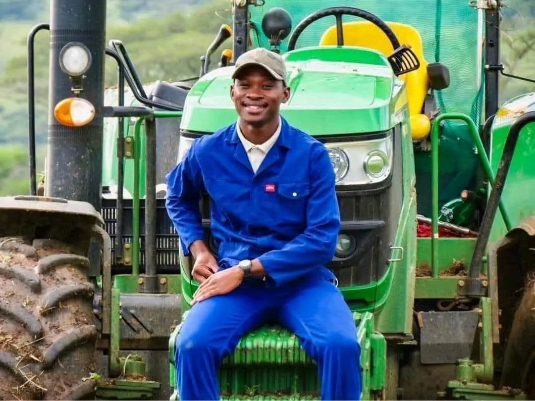 A smiling young man in blue overalls and a cap sits on a green tractor.