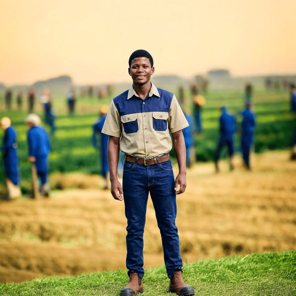 Smiling young man in a work shirt stands in a field with workers behind him.