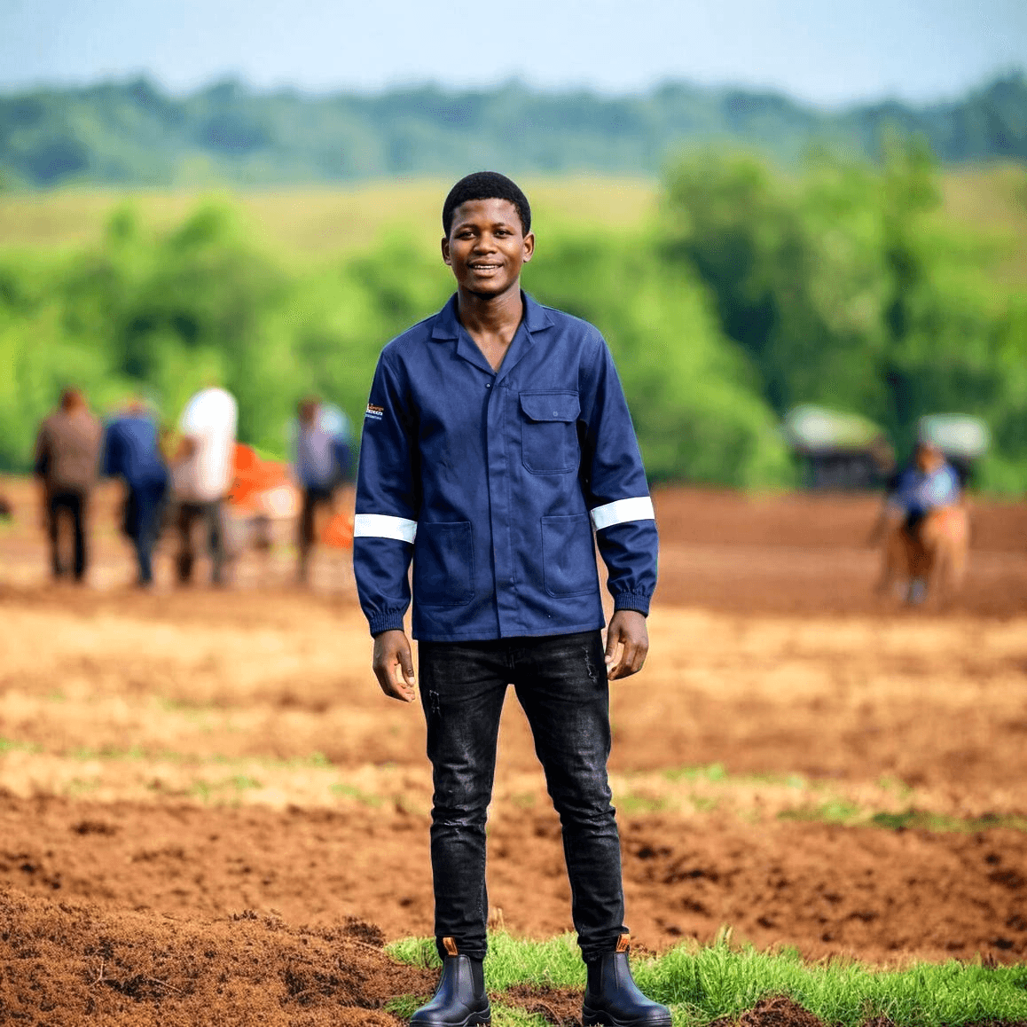 Smiling young man in a blue reflective work jacket stands in a tilled field.