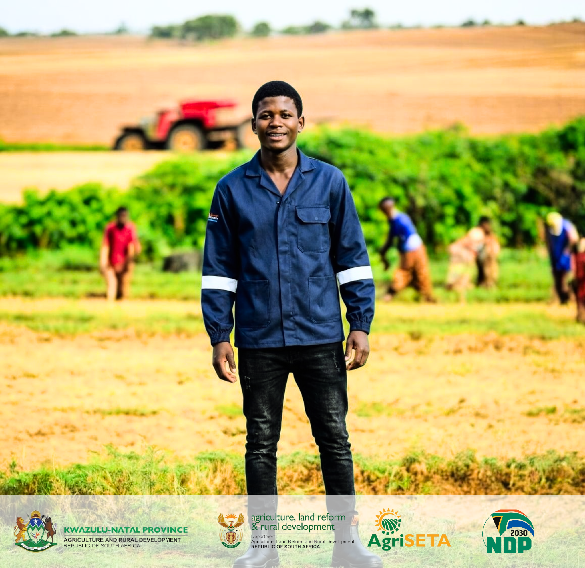Smiling young man in a blue work jacket standing in a sunny agricultural field.
